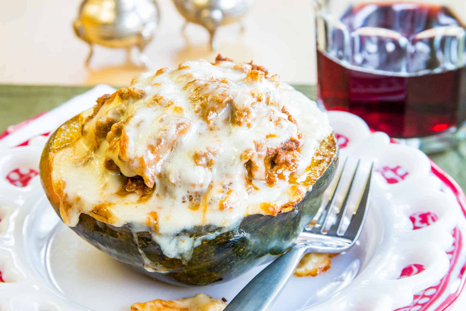 A stuffed acorn squash half filled with meat sauce and cheese on a plate next to a glass of wine.