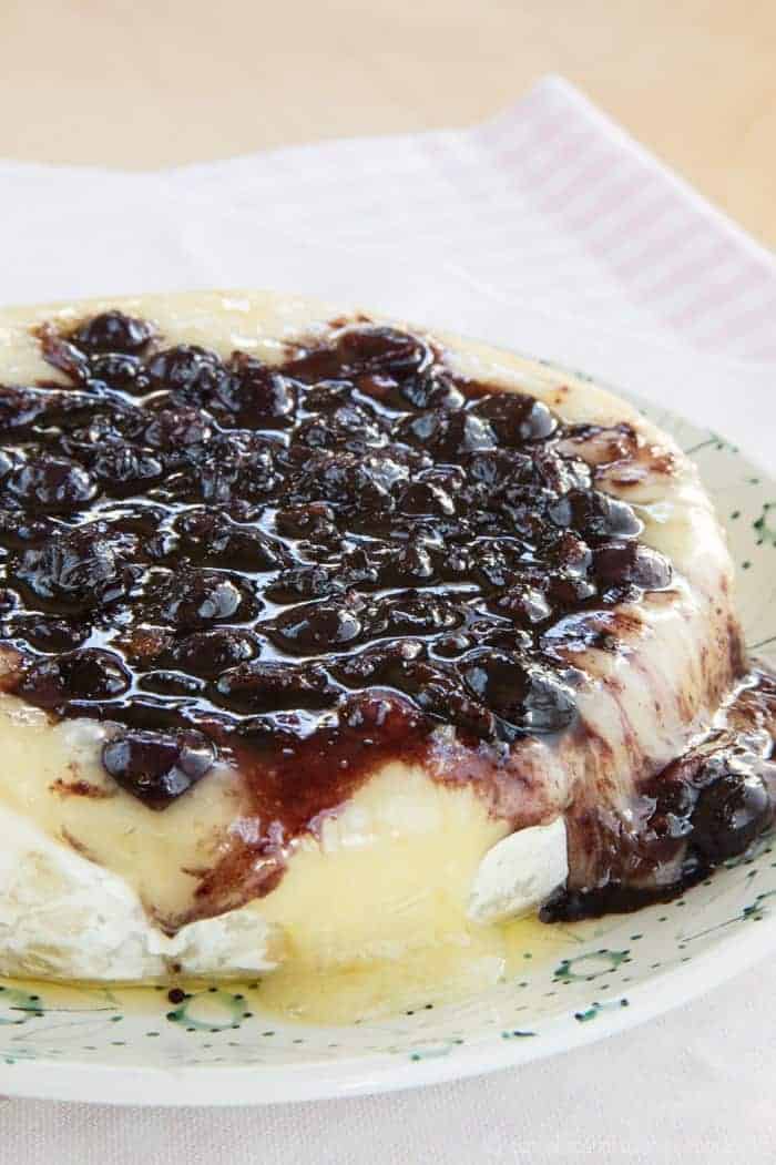 Melted baked Brie cheese topped with a balsamic blueberry and bacon jam, served on a plate.
