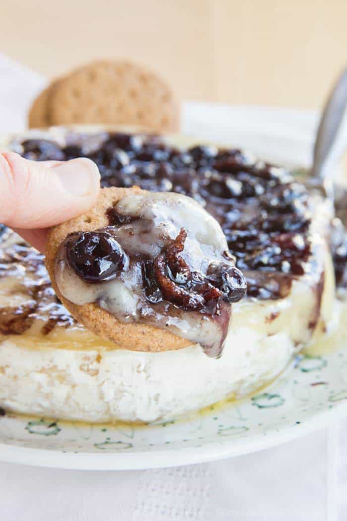 A cracker is dipped into baked brie and bacon balsamic blueberries.