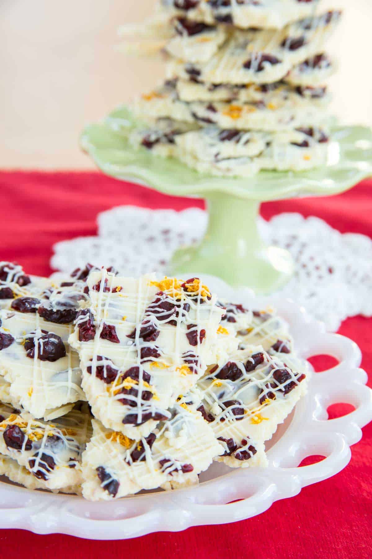 white chocolate bark with dried cranberries piled on a plate and a cake stand set on a red tablecloth.
