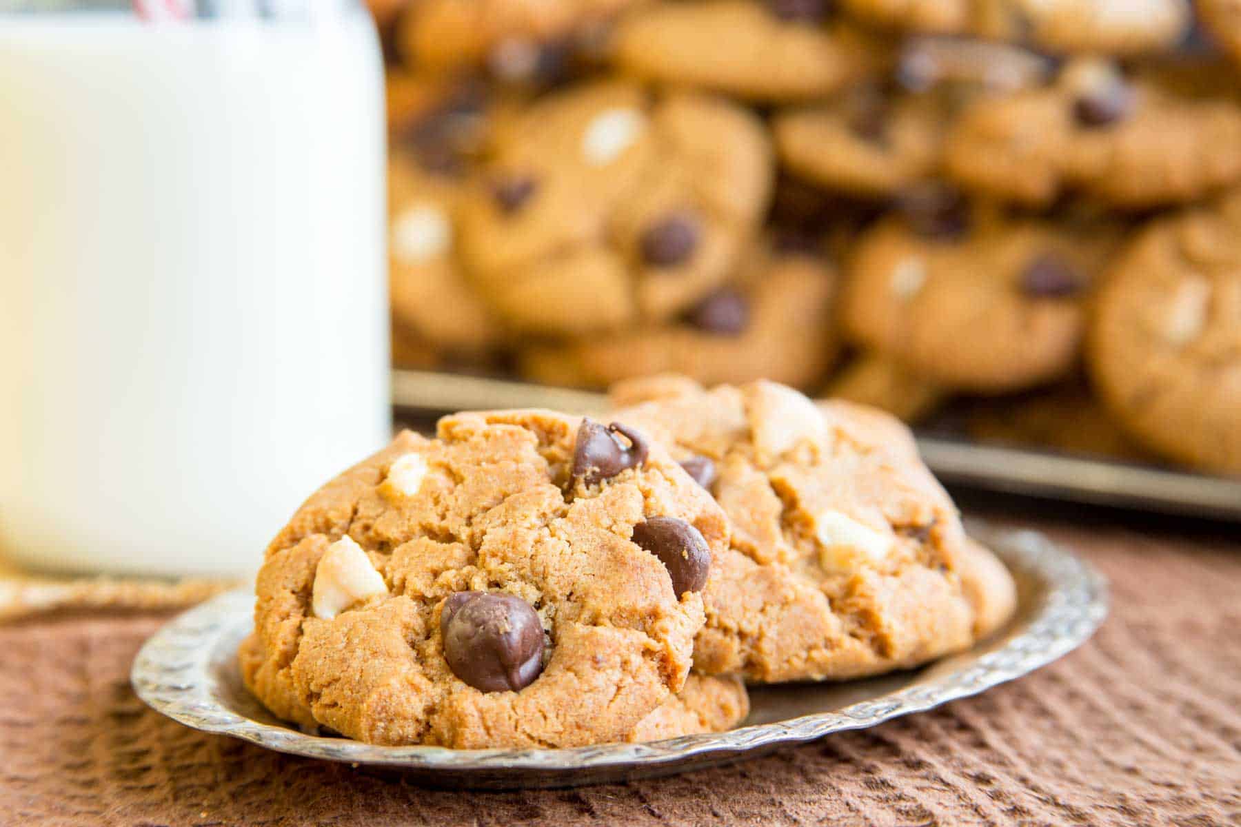 Three tripple chocolate chip peanut butter cookies on a small silver dish with a glass bottle of milk and more cookies piled in the background.