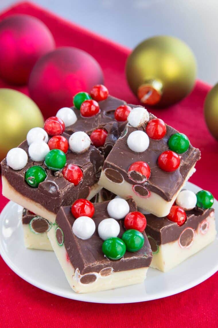 Two layer fudge with Christmas sixlet candies in it on a white plate on top of a red napkin with red and green ornaments in the background.