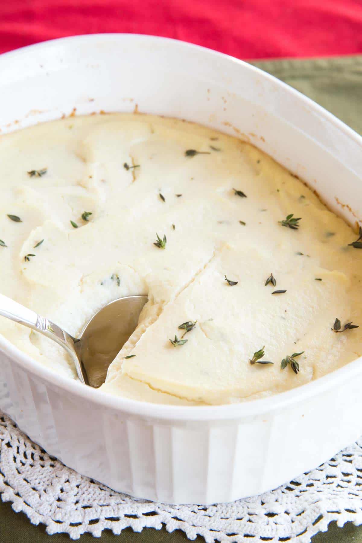 A serving spoon resting in a cauliflower casserole garnished with thyme on a white doily.