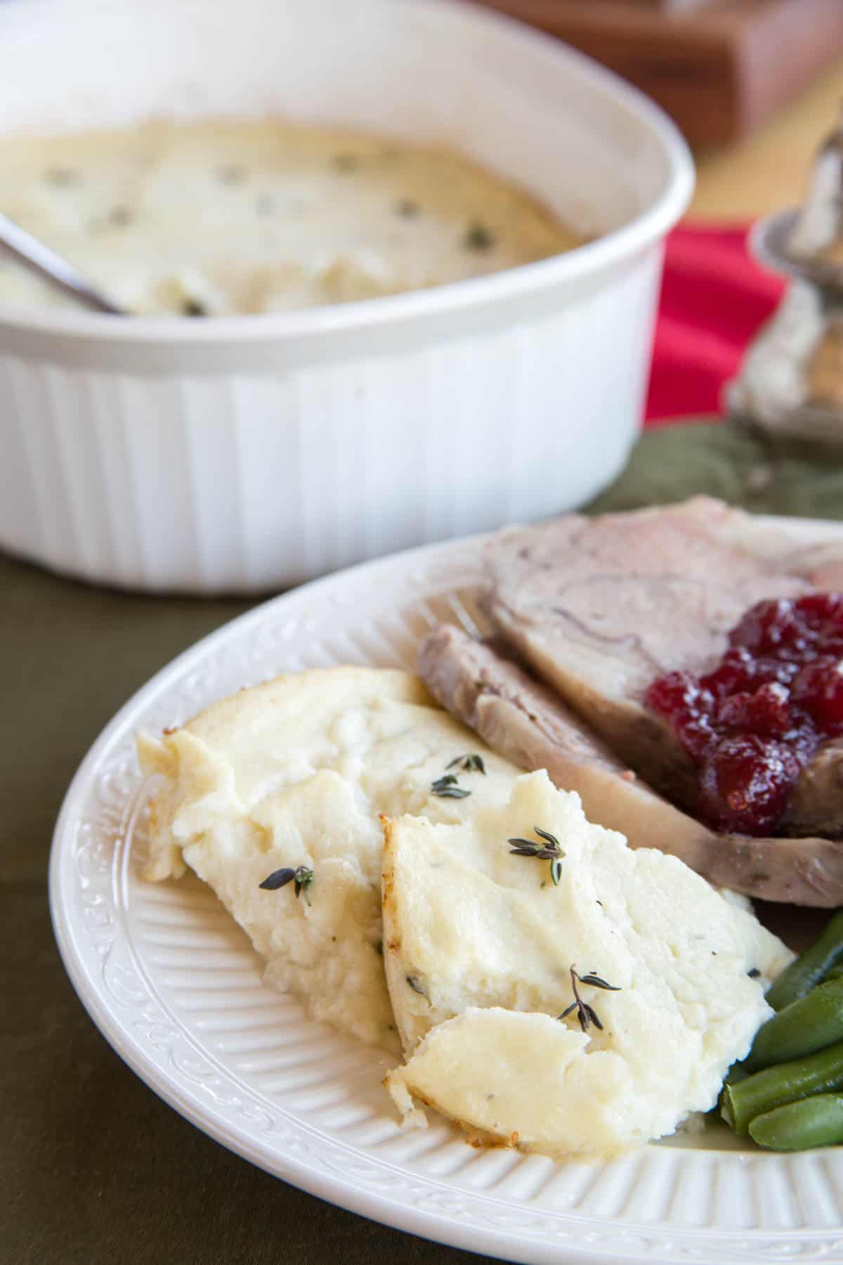 Goat cheese whipped cauliflower garnished with thyme served on a plate with green beans and sliced prime rib with the casserole dish in the background.