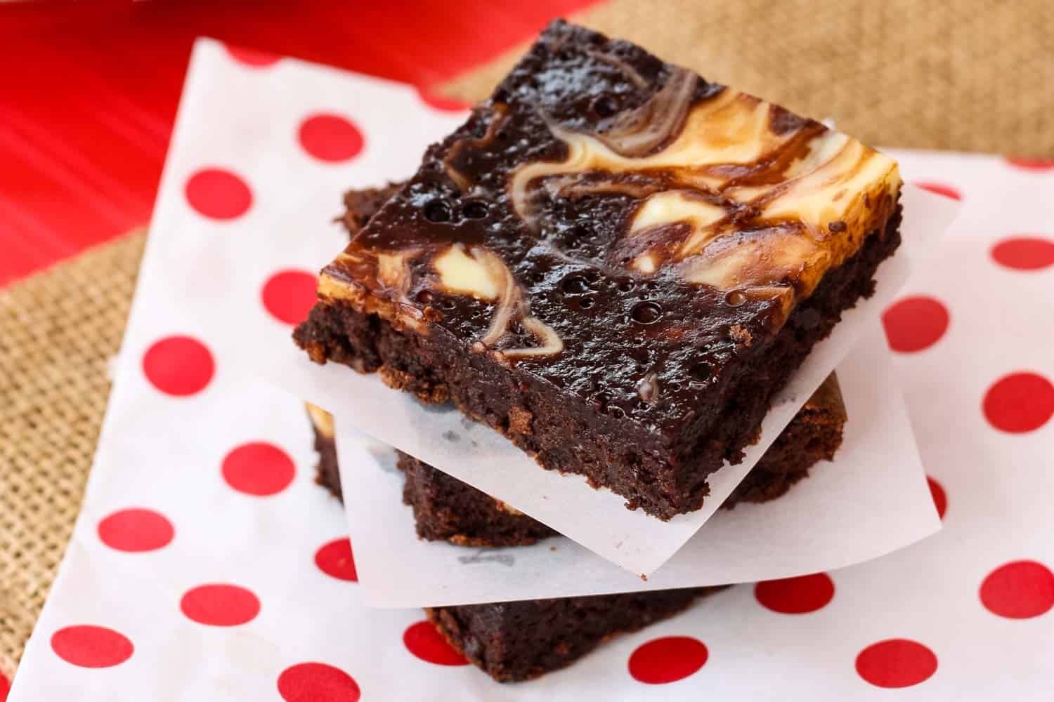Three gluten free brownies with swirls of cheesecake and nutella stacked on a napkin with red polka dots.