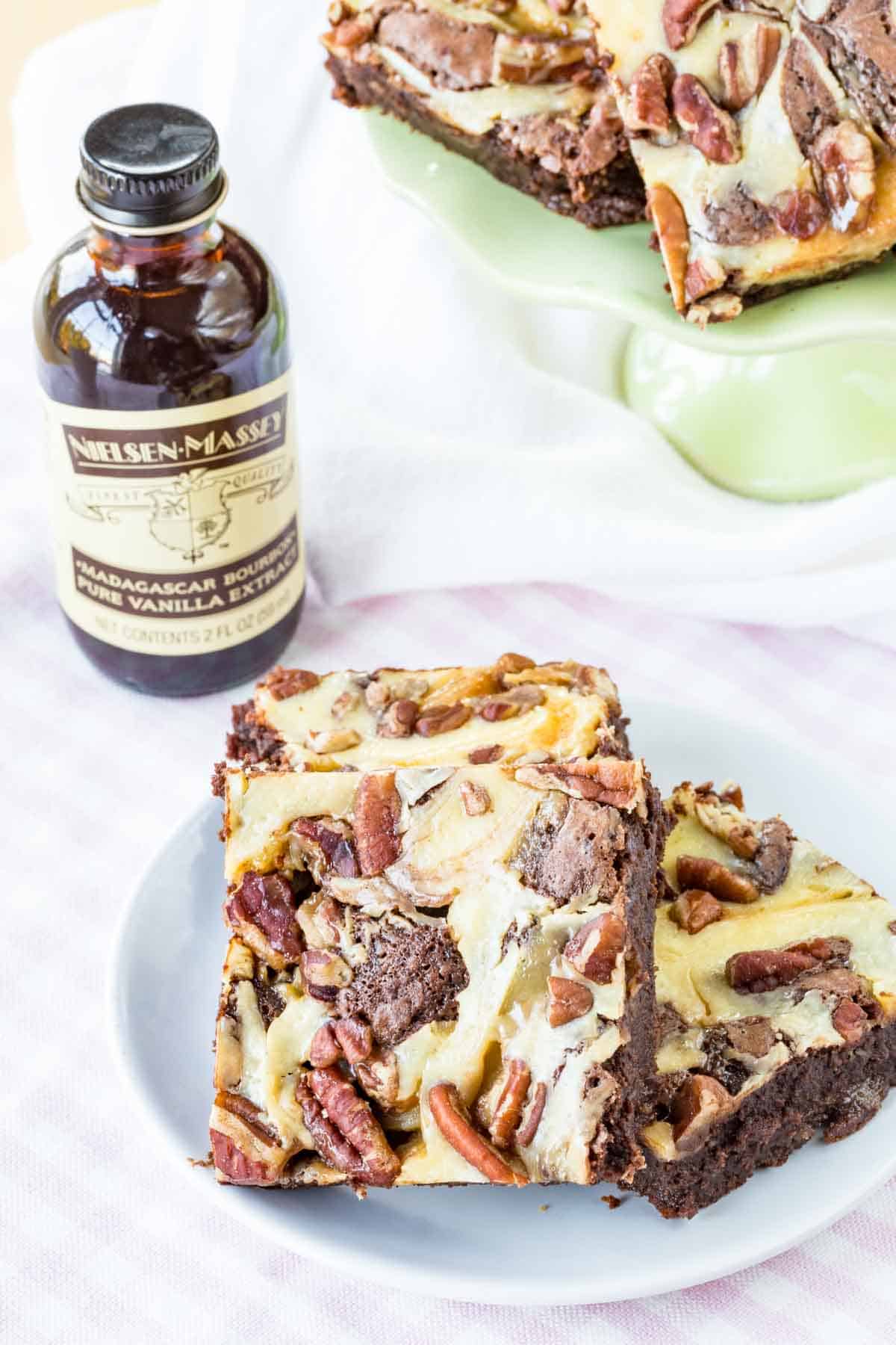 Three caramel pecan brownies on a small plate with a bottle of Nielsen-Massey vanilla extract next to them.