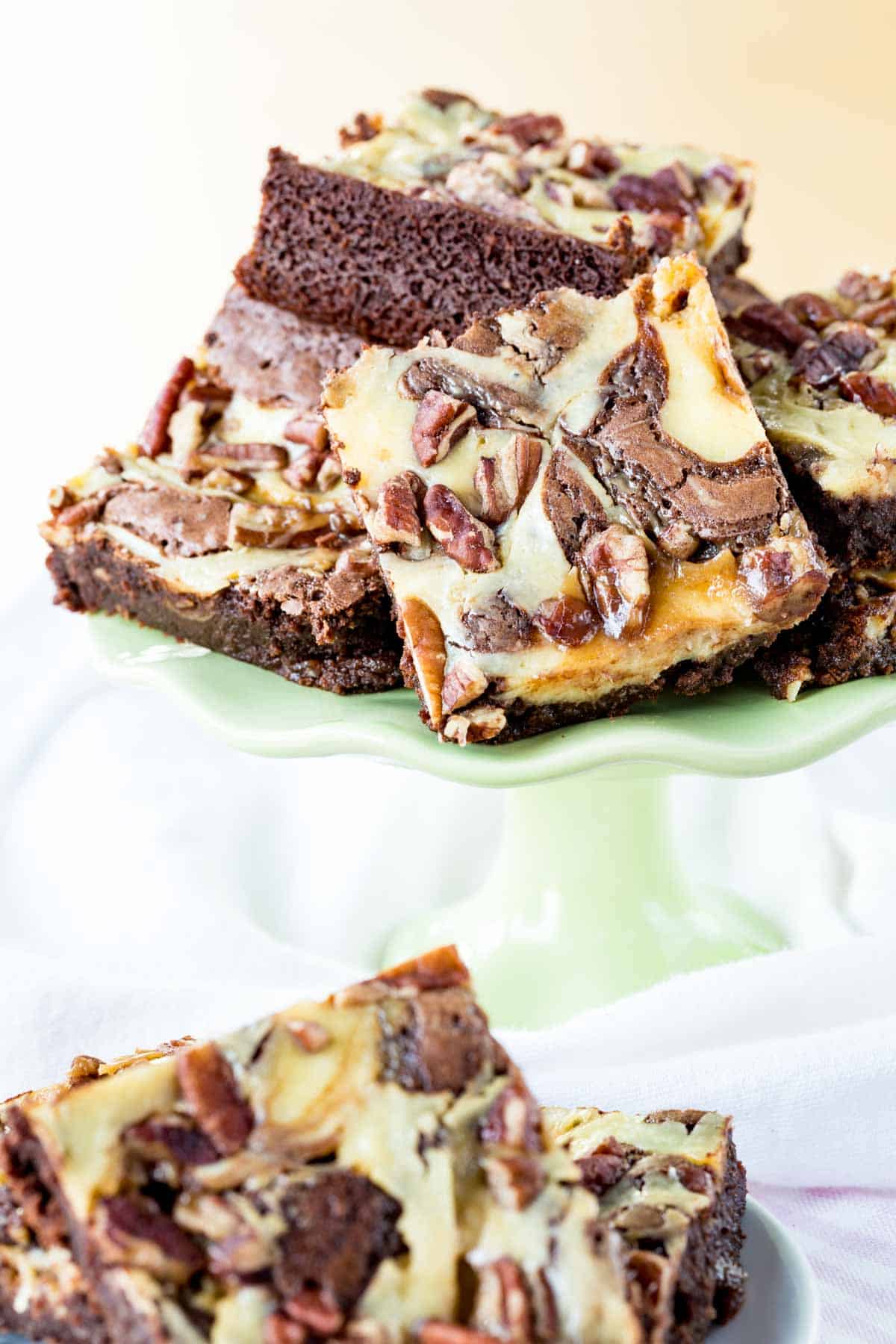 A pile of brownies topped with caramel, pecans, and cheesecake swirl on a green cake stand.