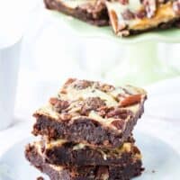 Three Flourless Turtle Cheesecake Brownies stacked on a plate with some on a cake stand in the background.