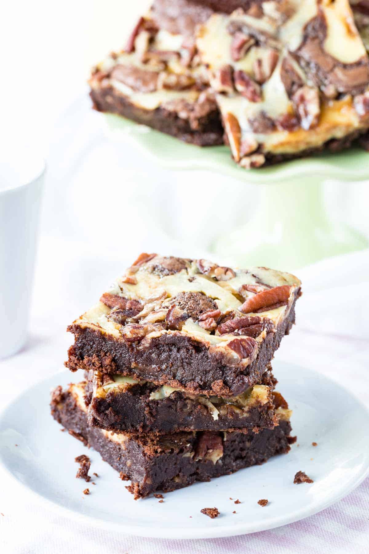 Three Flourless Turtle Cheesecake Brownies stacked on a plate with some on a cake stand in the background.