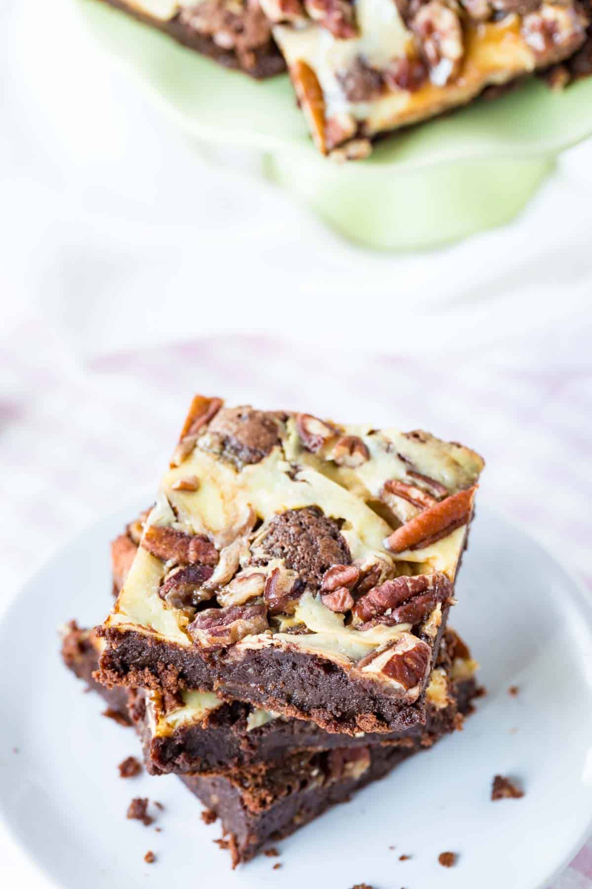 Three turtle brownies with a cheesecake swirl stacked on a white plate.