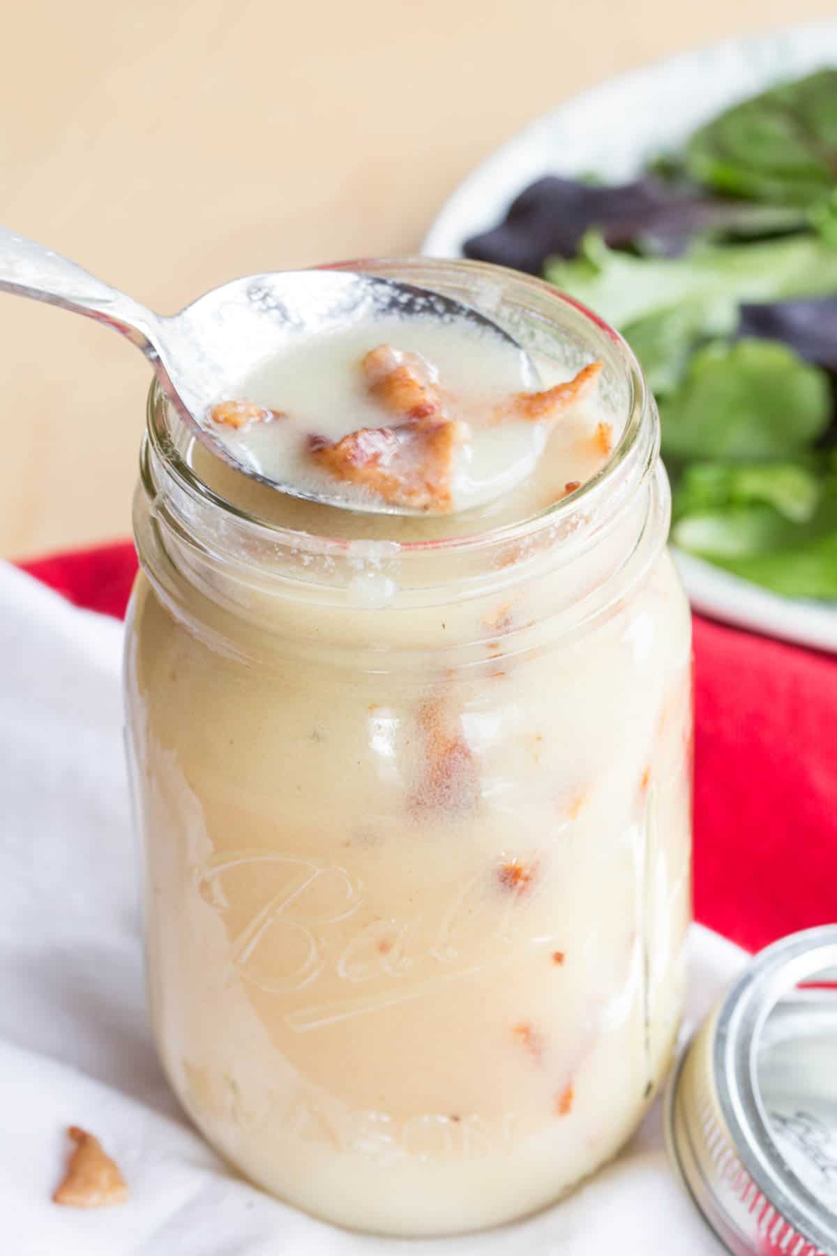 A spoon picking up some bacon dressing out of a full glass jar.