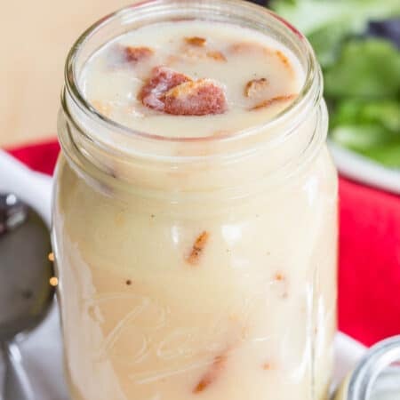 A glass jar filled with warm bacon dressing with a plate of lettuce behind it.