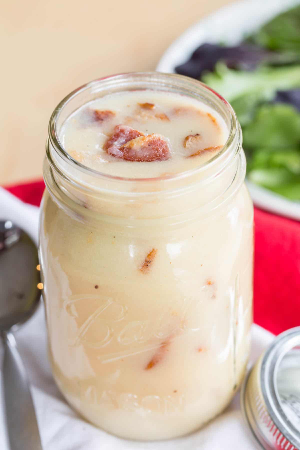 A glass jar filled with warm bacon dressing with a plate of lettuce behind it.