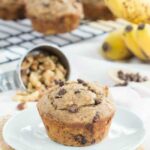 A browned butter banana chocolate chip muffin on a white plate, with muffins and bananas in the background.