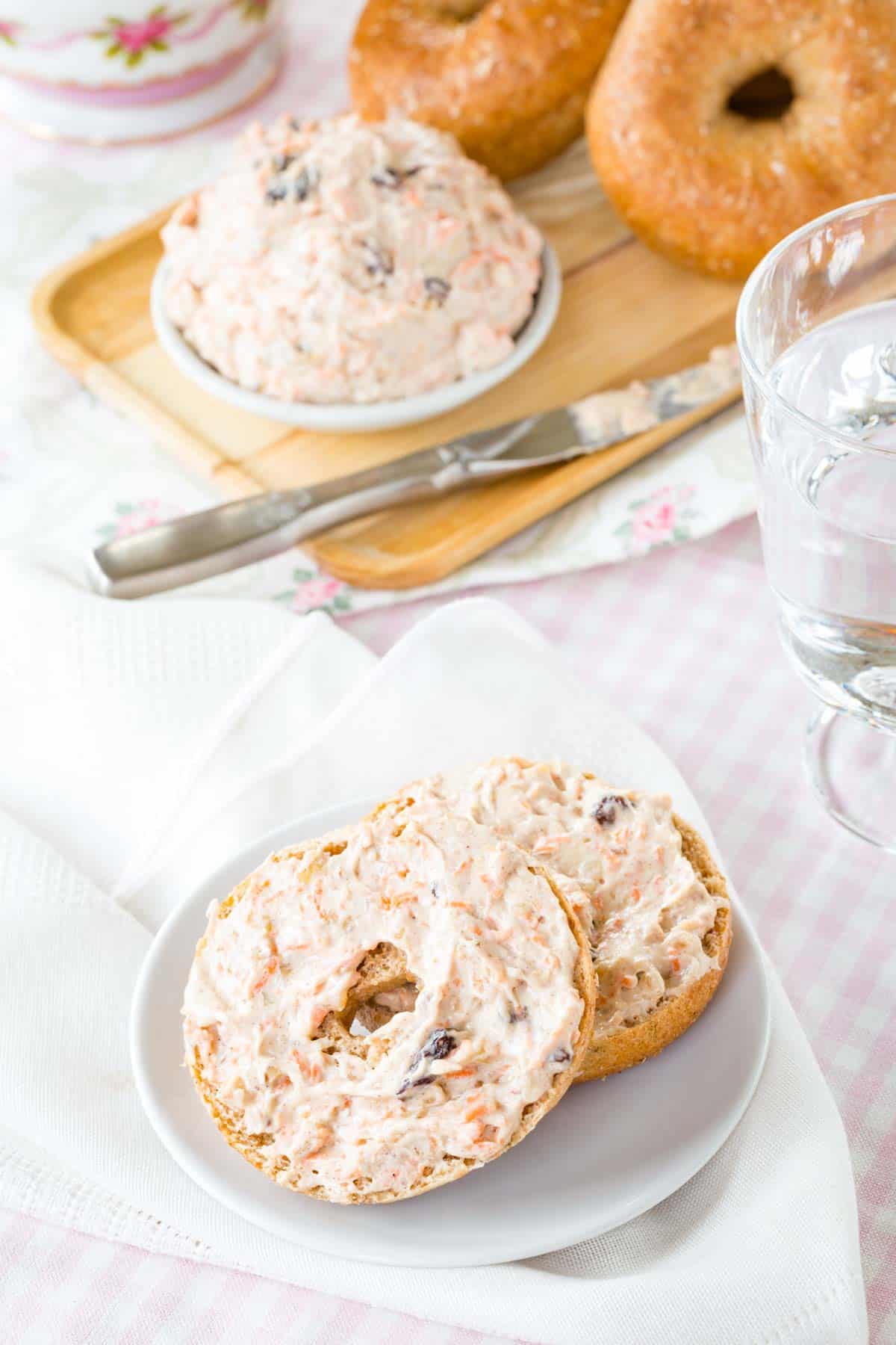 Two bagel hales covered with flavored cream cheese on a white plate on top of pink and white cloth napkins and a table cloth with platter of bagels and a bowl of cream cheese.