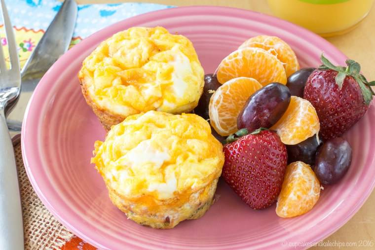 Two broccoli, ham and cheese egg muffins on a plate next to fruits.
