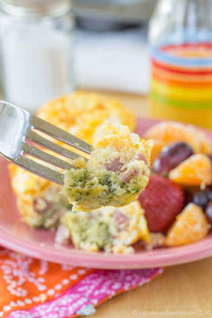 A fork cutting into a broccoli ham and cheese egg muffin on a pink plate next to fruit.