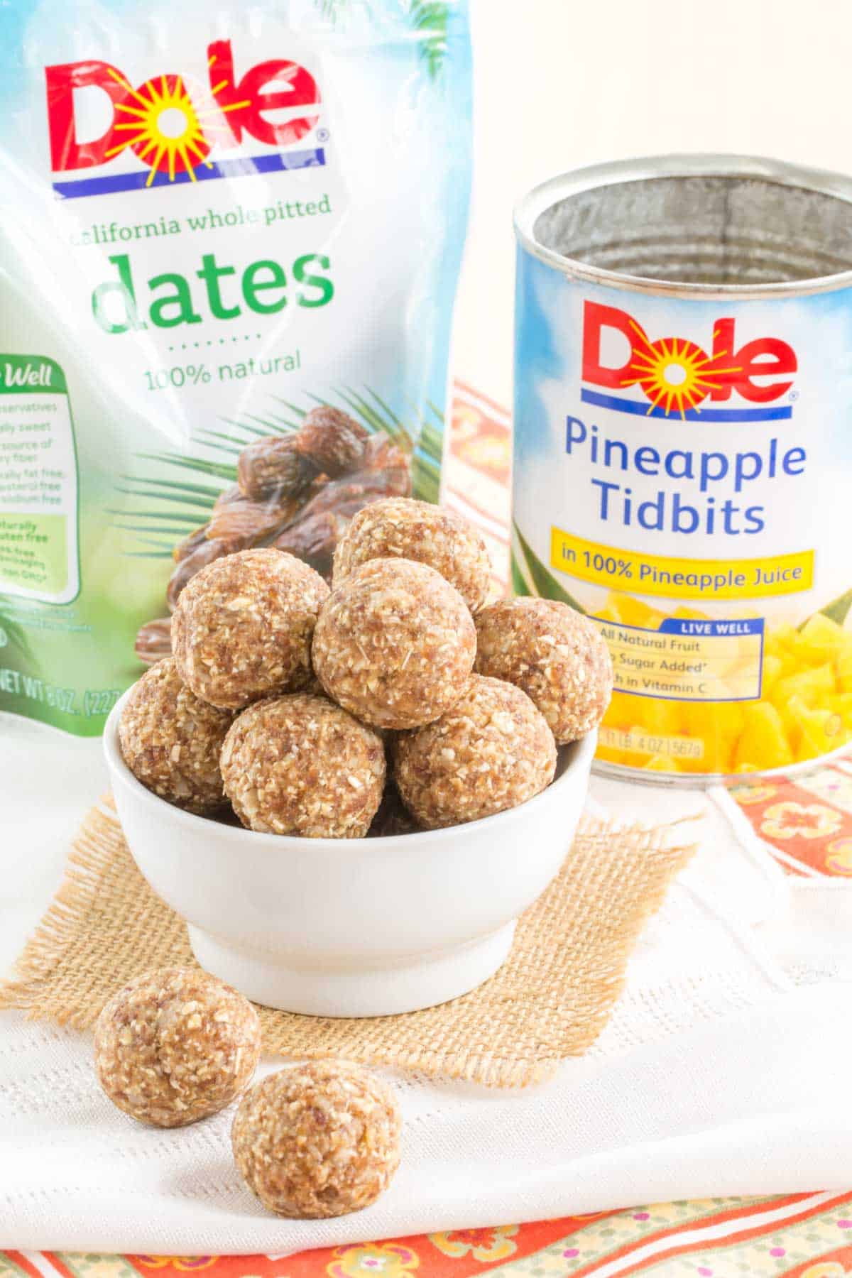 a bag of dates and a can of Dole pineapple tidbits behind a bowl of pineapple coconut energy balls with two on a napkin