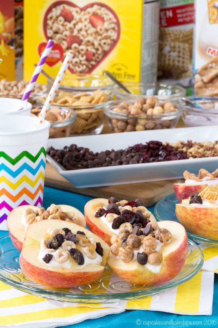 Yogurt and cereal stuffed apples on a plate with bowls and boxes of cereal around them.