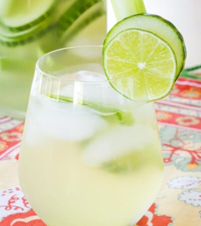 stemless wine glass filled with cucumber gimlet white sangria with more in a pitcher that has cucumber slices floating in it