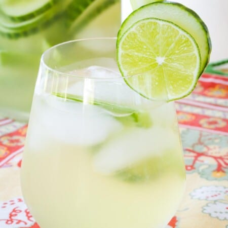 stemless wine glass filled with cucumber gimlet white sangria with more in a pitcher that has cucumber slices floating in it