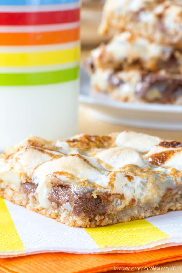 A s'mores bar on a white and yellow striped napkin with a plate of bars and a glass of milk in the background.