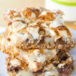 Three s'mores bars stacked on a plate with a glass of milk in the background.