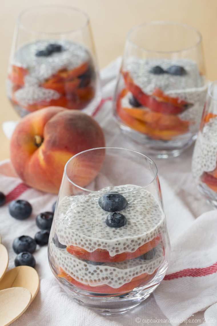 Blueberry and Peach Chia Pudding Parfaits in glasses, next to a whole peach and scattered blueberries.