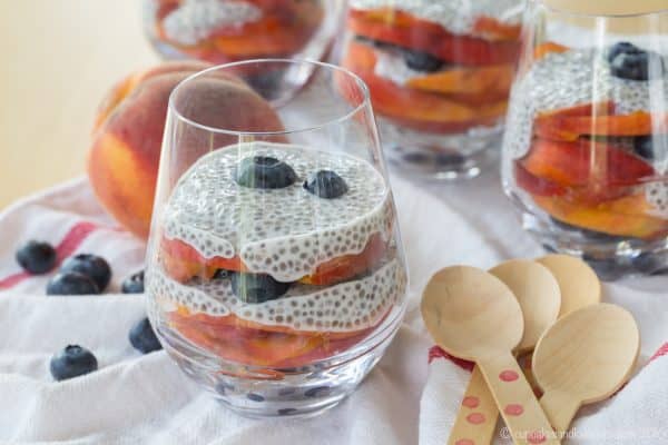 Blueberry and Peach Chia Pudding Parfaits in glasses, next to a whole peach and scattered blueberries.