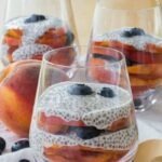 Blueberry and Peach Chia Pudding Parfaits in glasses, next to a whole peach and scattered blueberries.