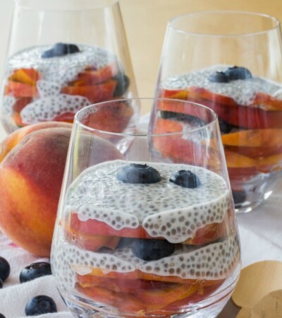 Blueberry and Peach Chia Pudding Parfaits in glasses, next to a whole peach and scattered blueberries.