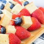 Mini fruit kabobs each with a blueberry, a piece of pound cake, and a strawberry on a toothpick set out on a wooden platter.