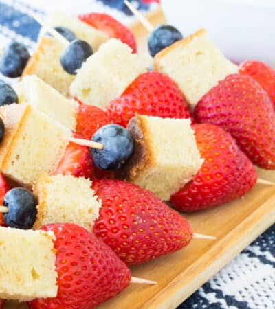 Mini fruit kabobs each with a blueberry, a piece of pound cake, and a strawberry on a toothpick set out on a wooden platter.