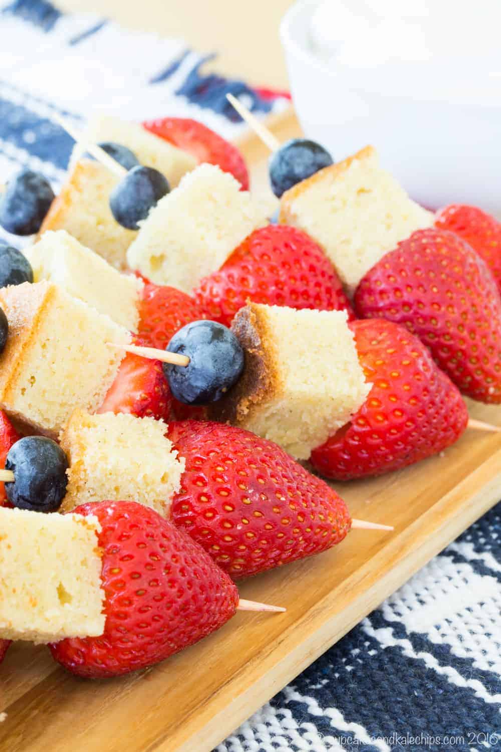 Mini fruit kabobs each with a blueberry, a piece of pound cake, and a strawberry on a toothpick set out on a wooden platter.