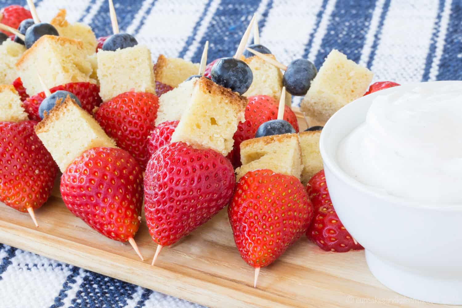 Mini fruit kabobs with pound cake cubes on a wooden platter next to a bowl of whipped cream.