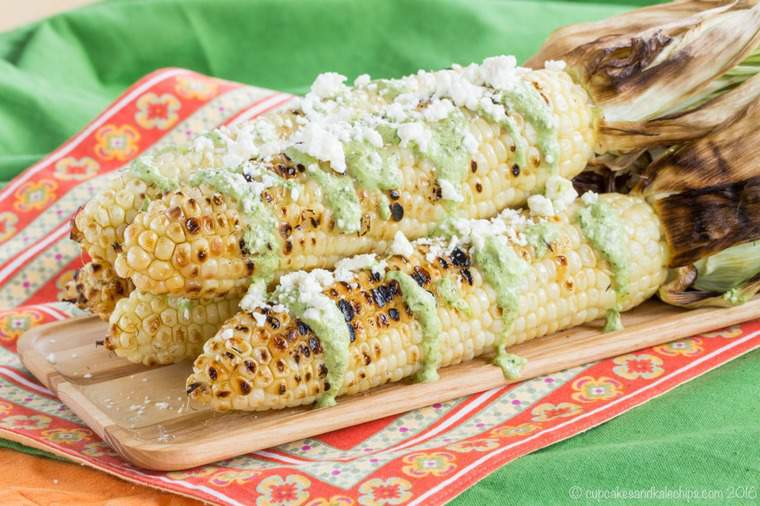 ears of grilled corn stacked ona  wooden platter drizzled with a green sauce and topped with crumbled cheese.