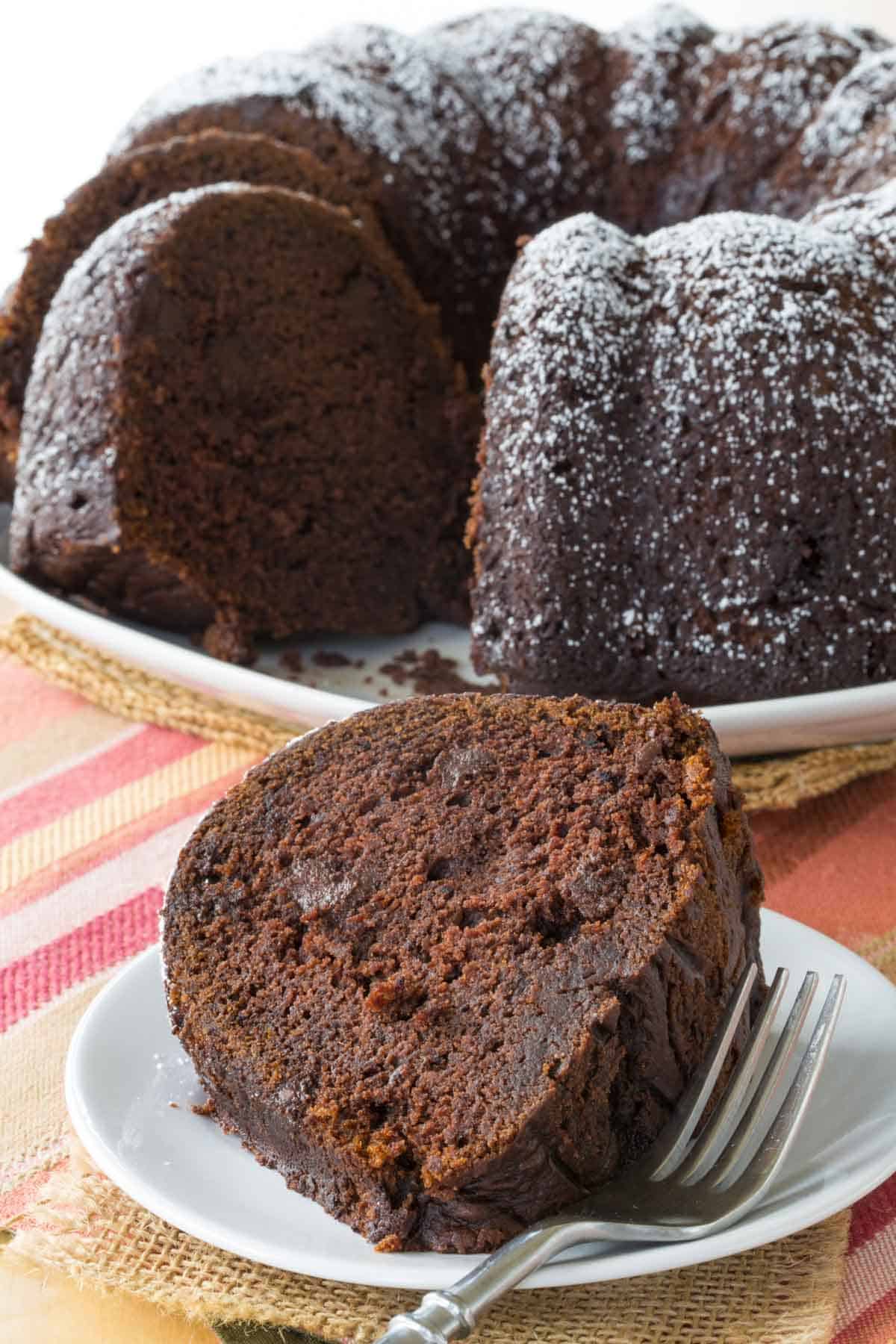 piece Gluten-Free Chocolate Pumpkin Spice Cake on a white plate in front of the rest of the cake on a striped placemat