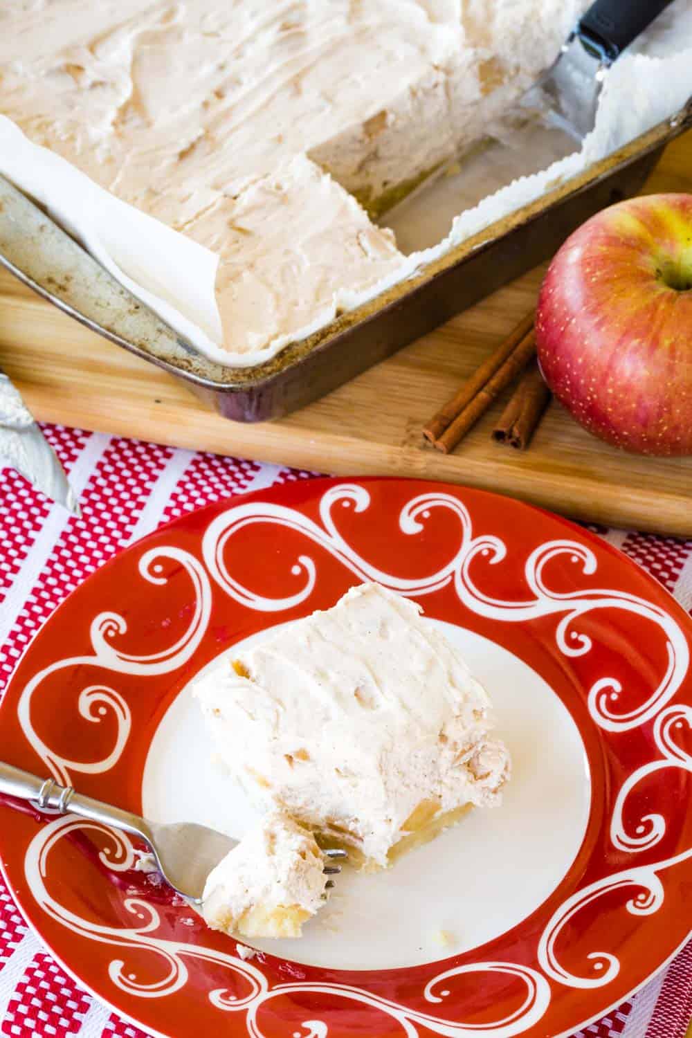 A slice of no-bake apple cheesecake bars on a red and white plate with the pan with the piece removed on a wooden board next to the plate.