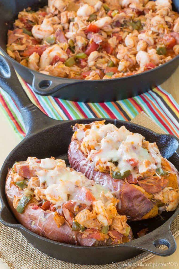 Two baked sweet potatoes in a cast iron skillet stuffed with a jambalaya mixture and topped with melted cheese.