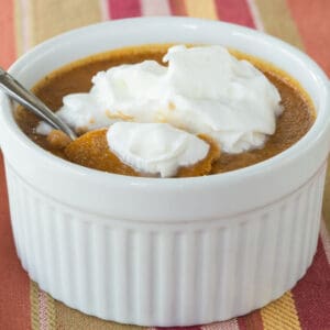 A spoon picking up a bite of a crustless pumpkin pie baked in a ramekin with whipped cream on top.