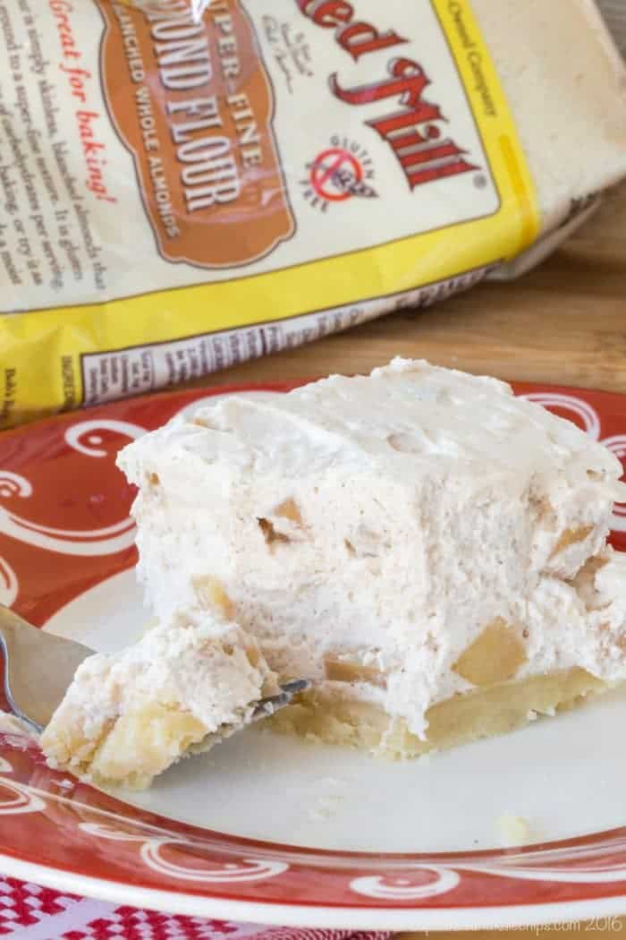 A closeup of an apple cheesecake bar on a plate with a bag of almond flour behind it.