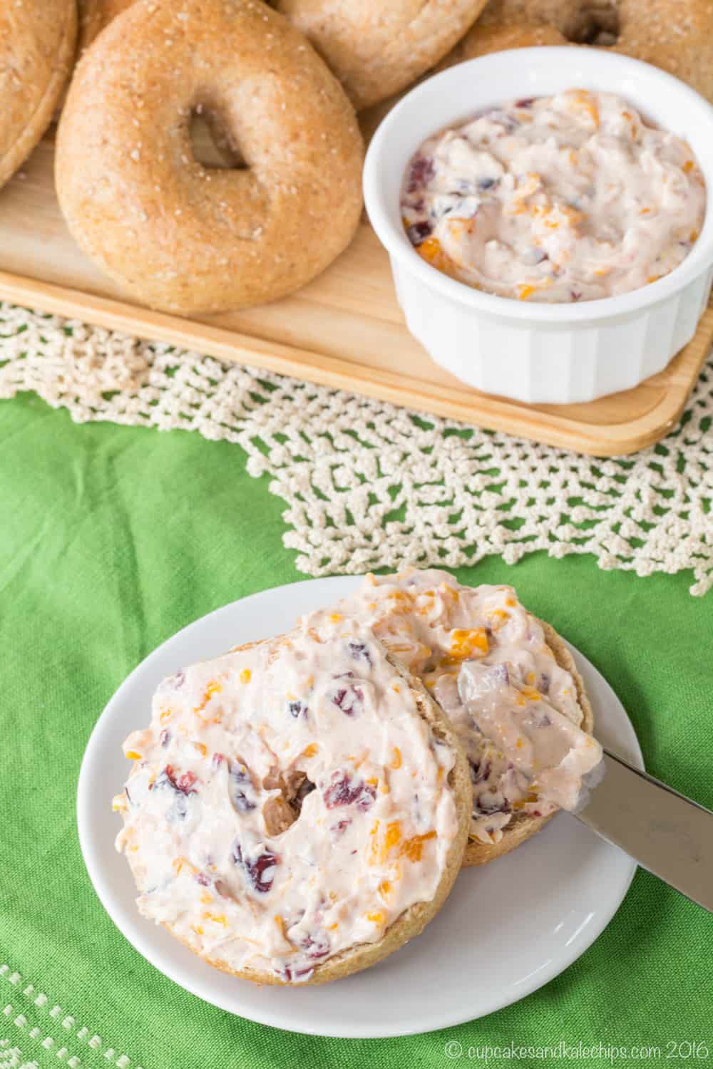 Looking down at a table covered with a green table cloth with a wooden platter of bagels and flavored cream cheese and one of the bagels on a plate spread with orange cranberry cream cheese.