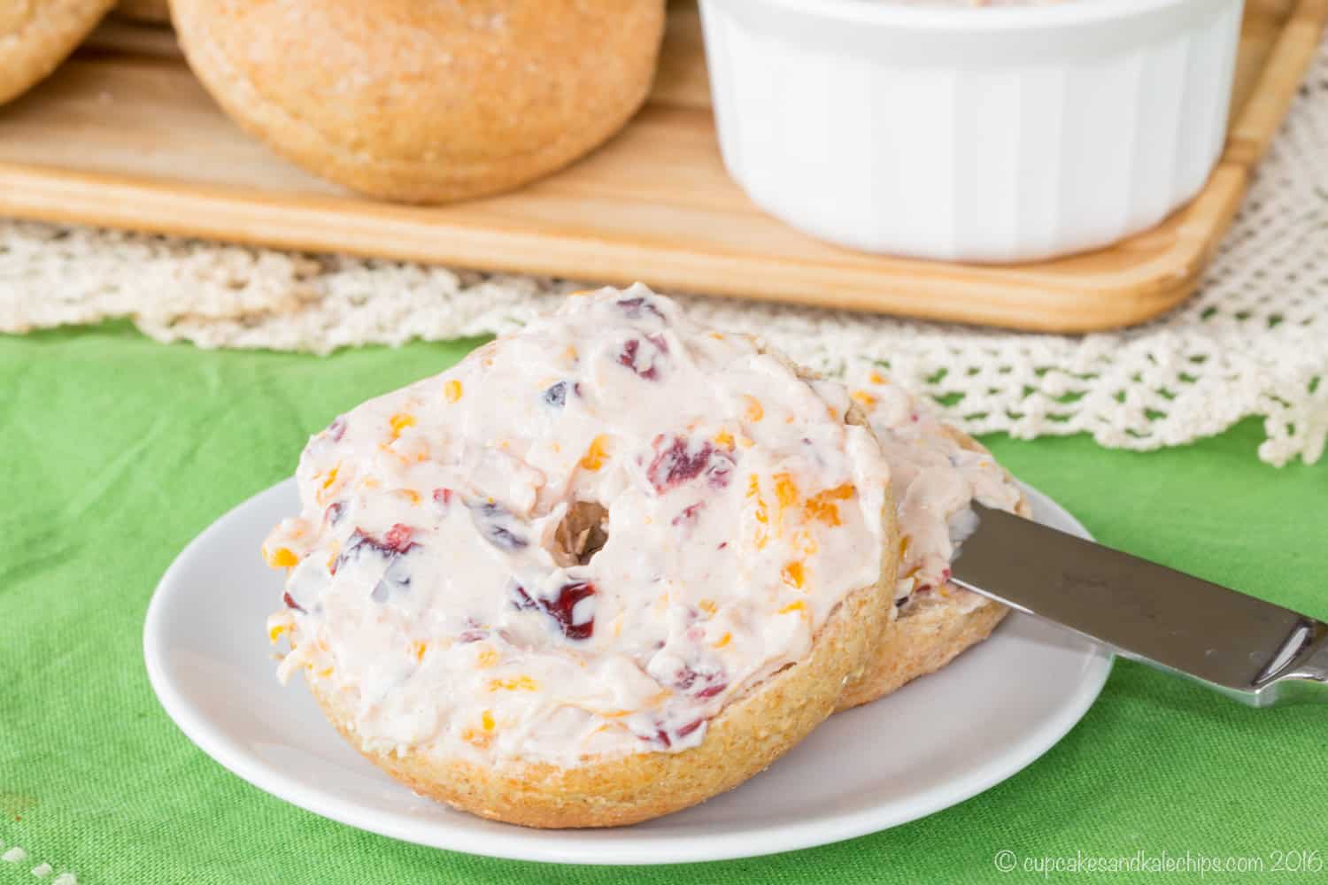 A mini whole wheat bagel covered with orange cranberry cream cheese on a white plate on a green tablecloth.