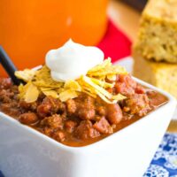 white bowl of beef chili with beans topped with crushed tortilla chips, shredded cheese, and a dollop of sour cream