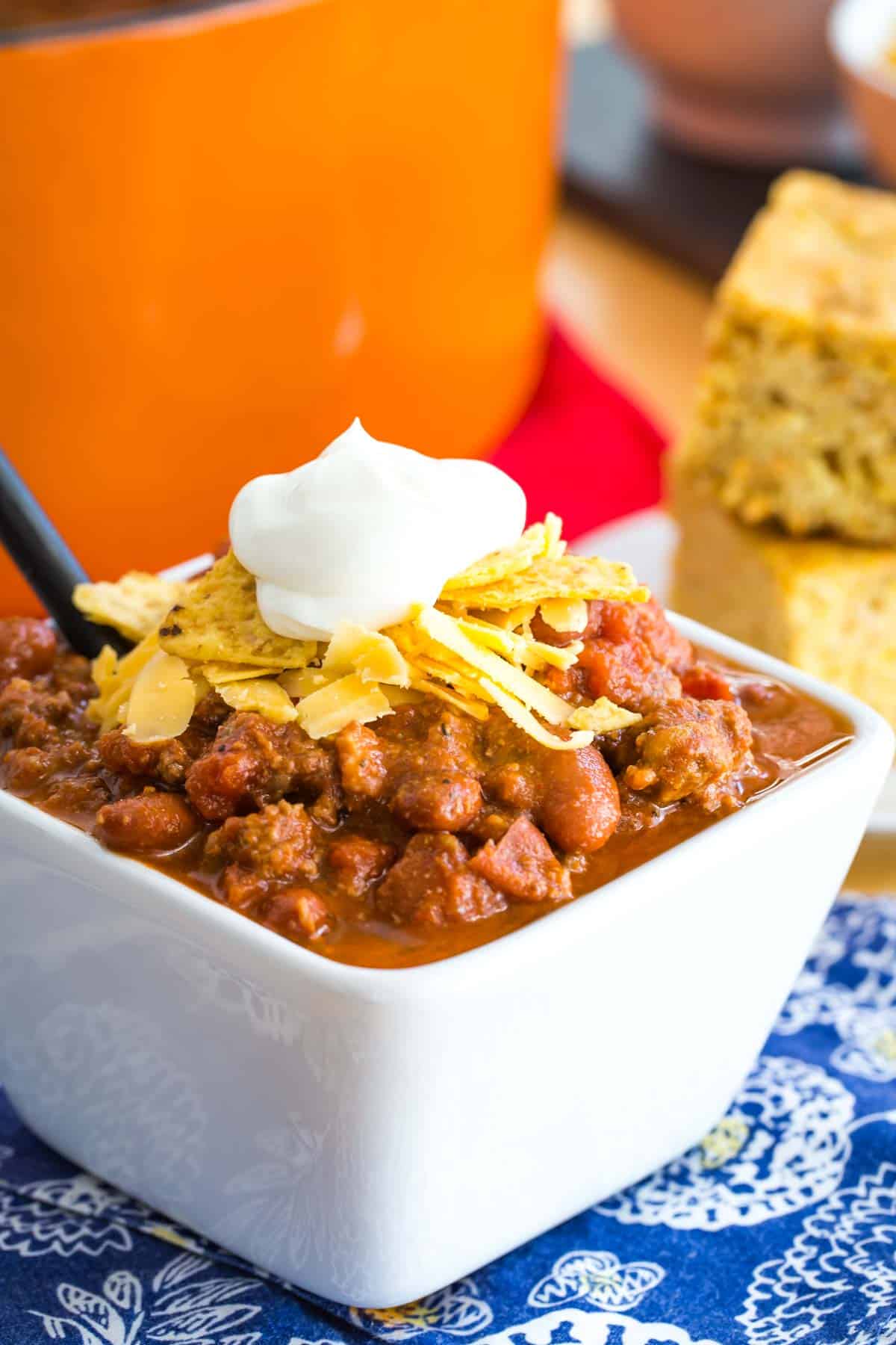 white bowl of beef chili with beans topped with crushed tortilla chips, shredded cheese, and a dollop of sour cream