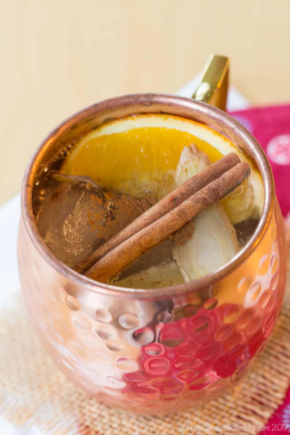 Looking down into a cold toddy whiskey mule cocktail in a copper mug on a piece of burlap with orange, lemon, cinnamon stick, and a slice of fresh ginger as garnishes.