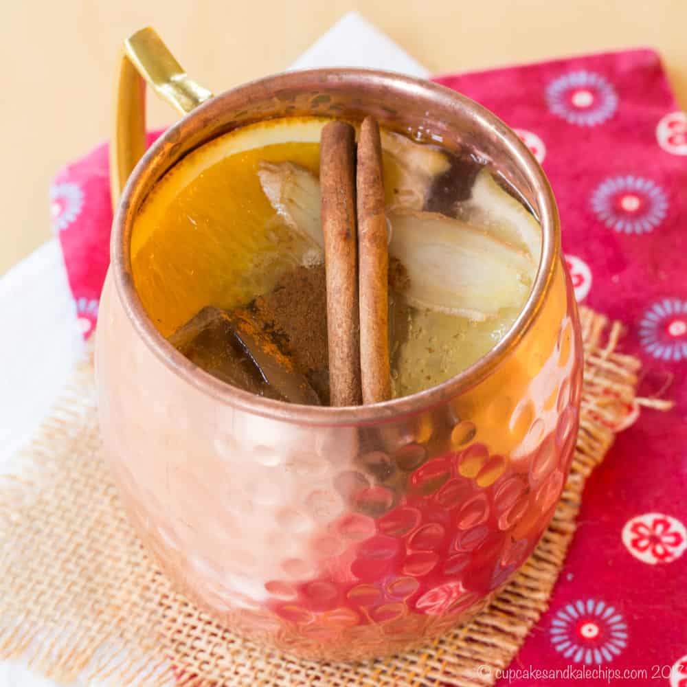 A whiskey mule cocktail in a copper mug with an orange slice and cinnamon stick on top of a red cloth napkin and piece of burlap.