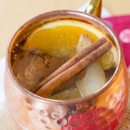 Looking down into a copped much with a cocktail with floating orange and lemon slices, and a cinnamon stick and text overlay that says "Cold Toddy Whiskey Mule".