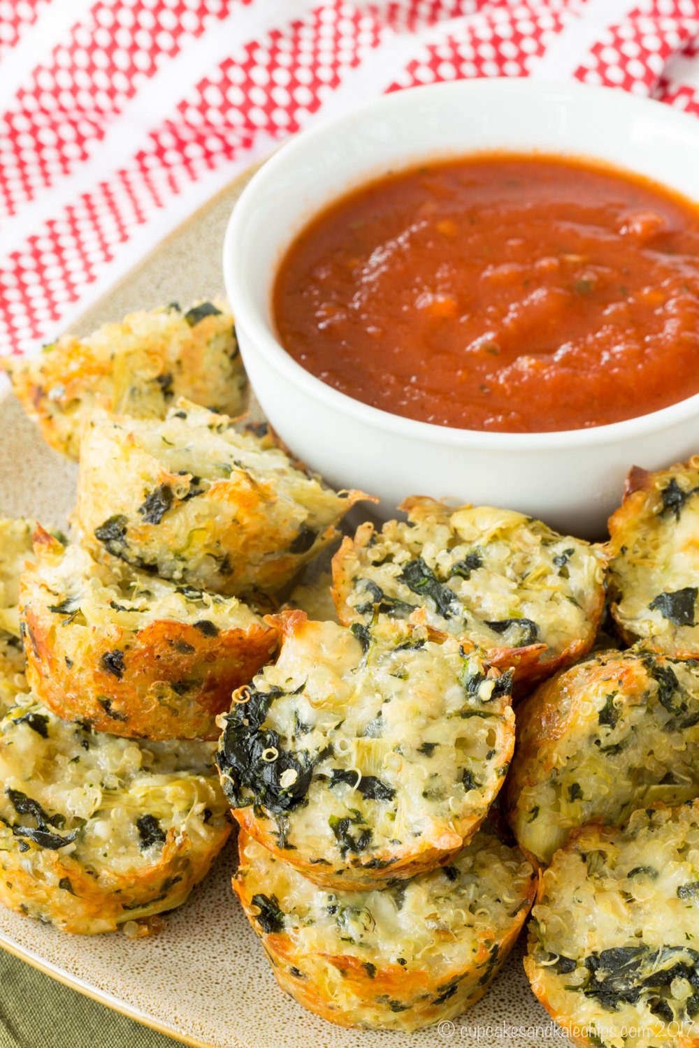 A platter of spinach artichoke bites made with quinoa on a plate with a bowl of tomato sauce.