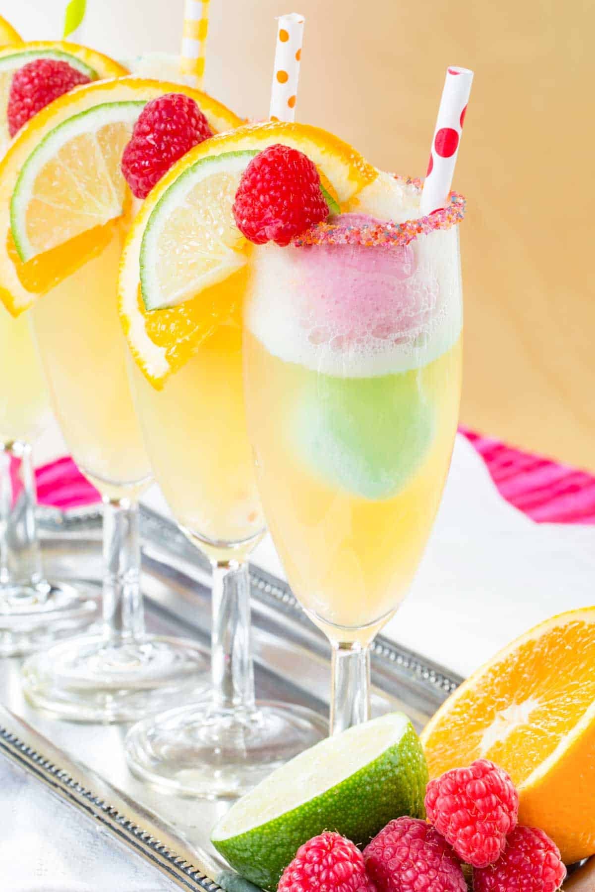 Festively decorated flutes filled with rainbow sherbet floats on a silver tray with fruit on it..